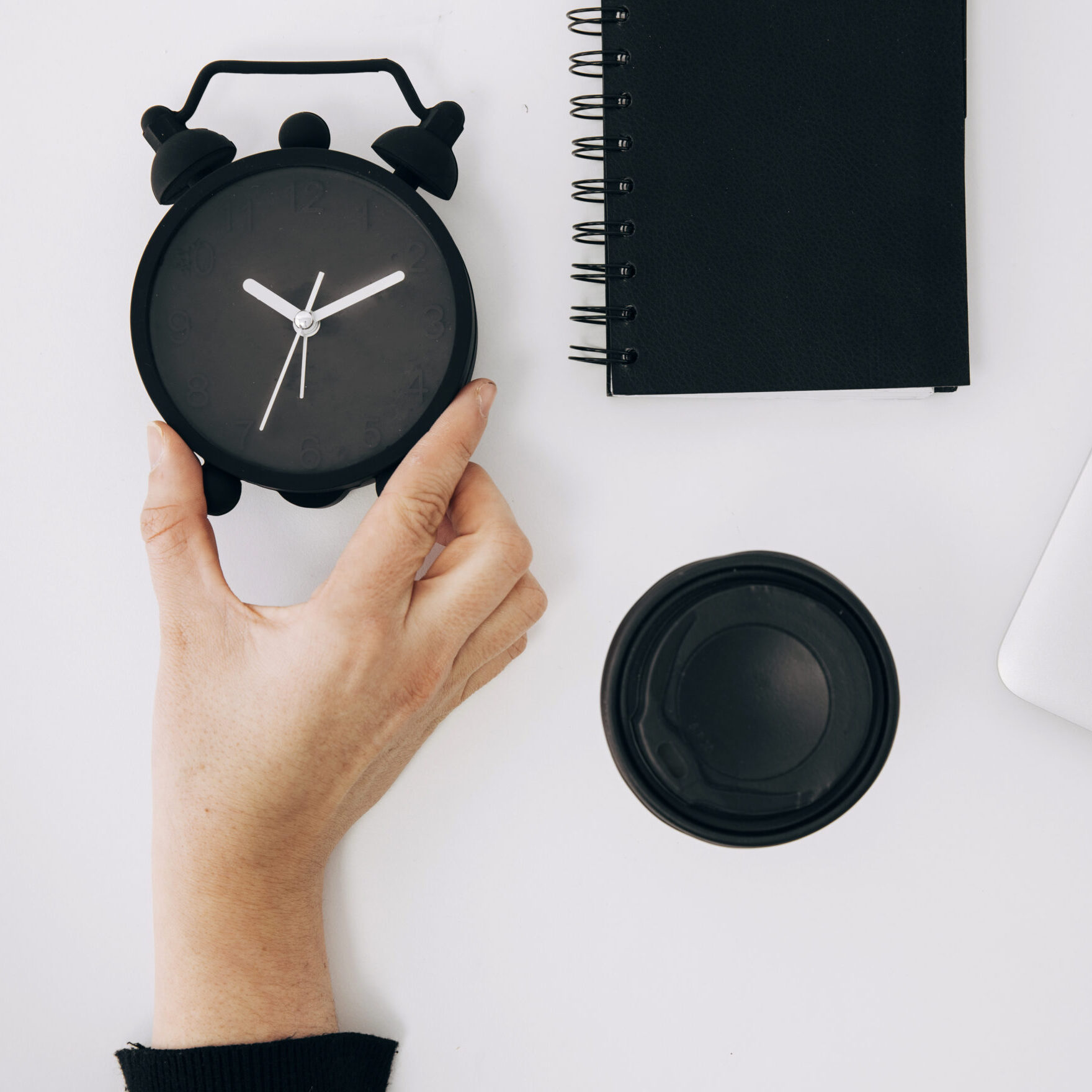 Home close up person s hand holding black alarm clock spiral notepad laptop takeaway coffee cup white desk